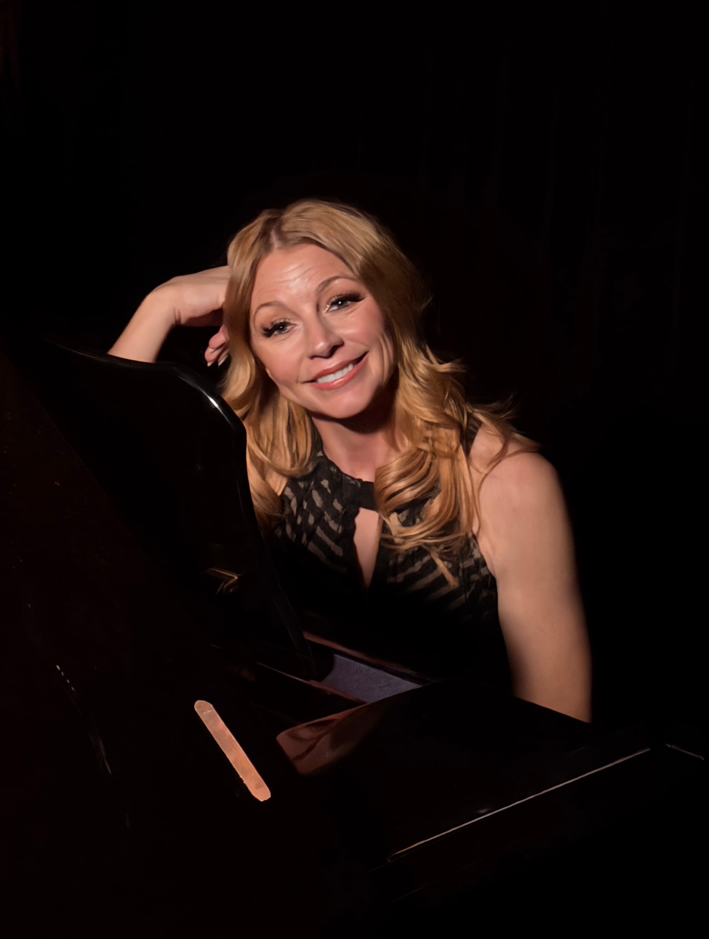 Lisa Apple at the piano — relaxed, warm smile in dramatic lighting